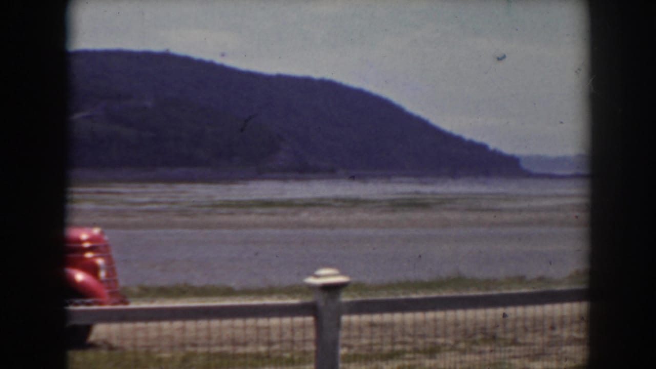 Red car partly visible beside a wooden fence overlooking a shallow riverbed and distant hills.