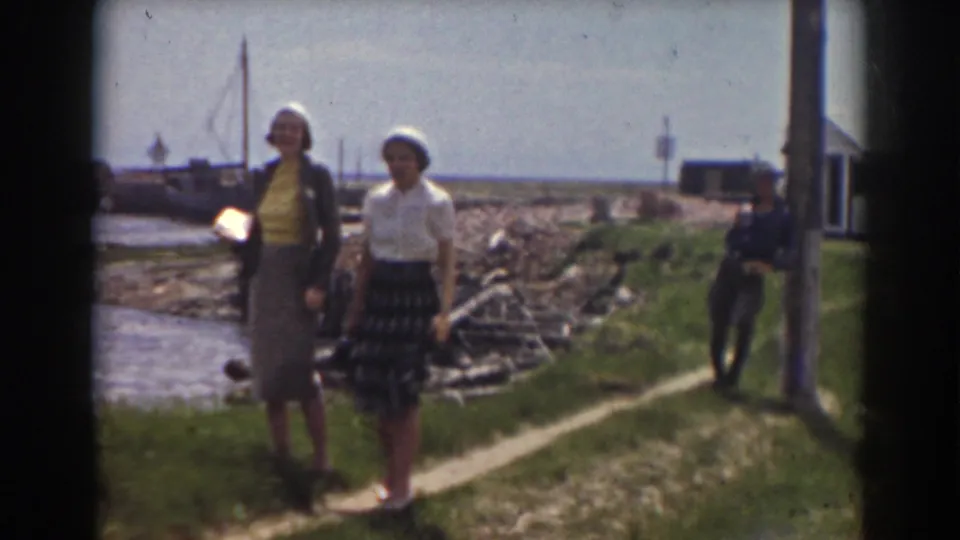 Two women face the camera on a grassy waterfront path, with boats and shoreline structures behind them.