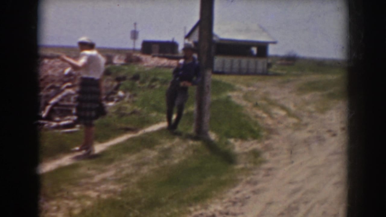 Two women and a man stand near a dirt path, grassy bank, wooden pole, and small building.