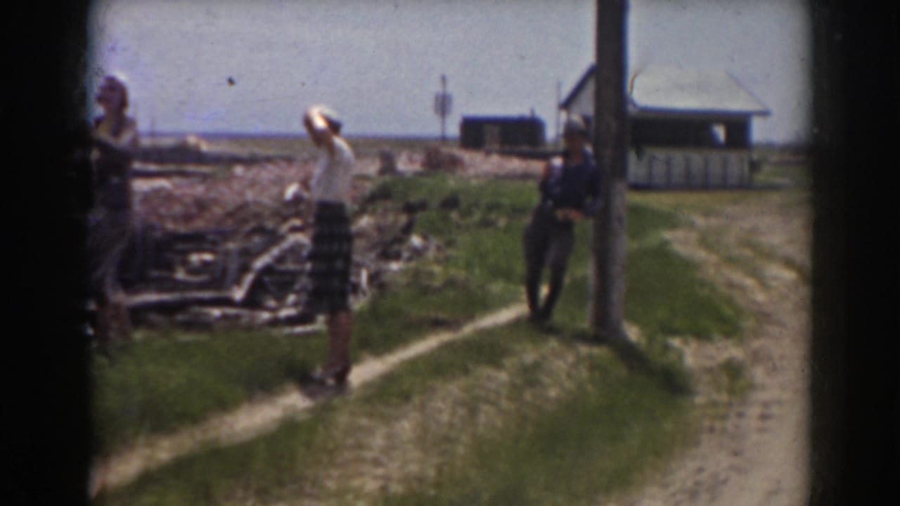A woman in a light blouse and patterned skirt stands near piled material as a man in a hat leans by a pole in the 1938 New Hampshire scene.