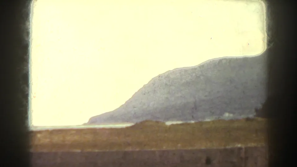 The frame shows a distant mountain slope or large hillside across water, with heavy exposure and film-gate edges from the 8mm home movie.