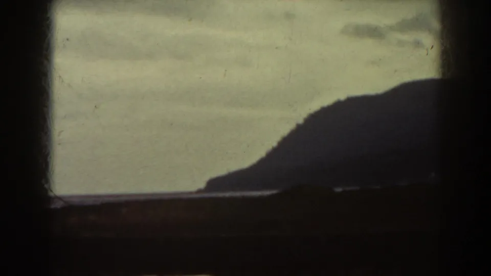 A dark mountain slope is seen across water under a muted sky, framed by the visible edges of the original 8mm film image.