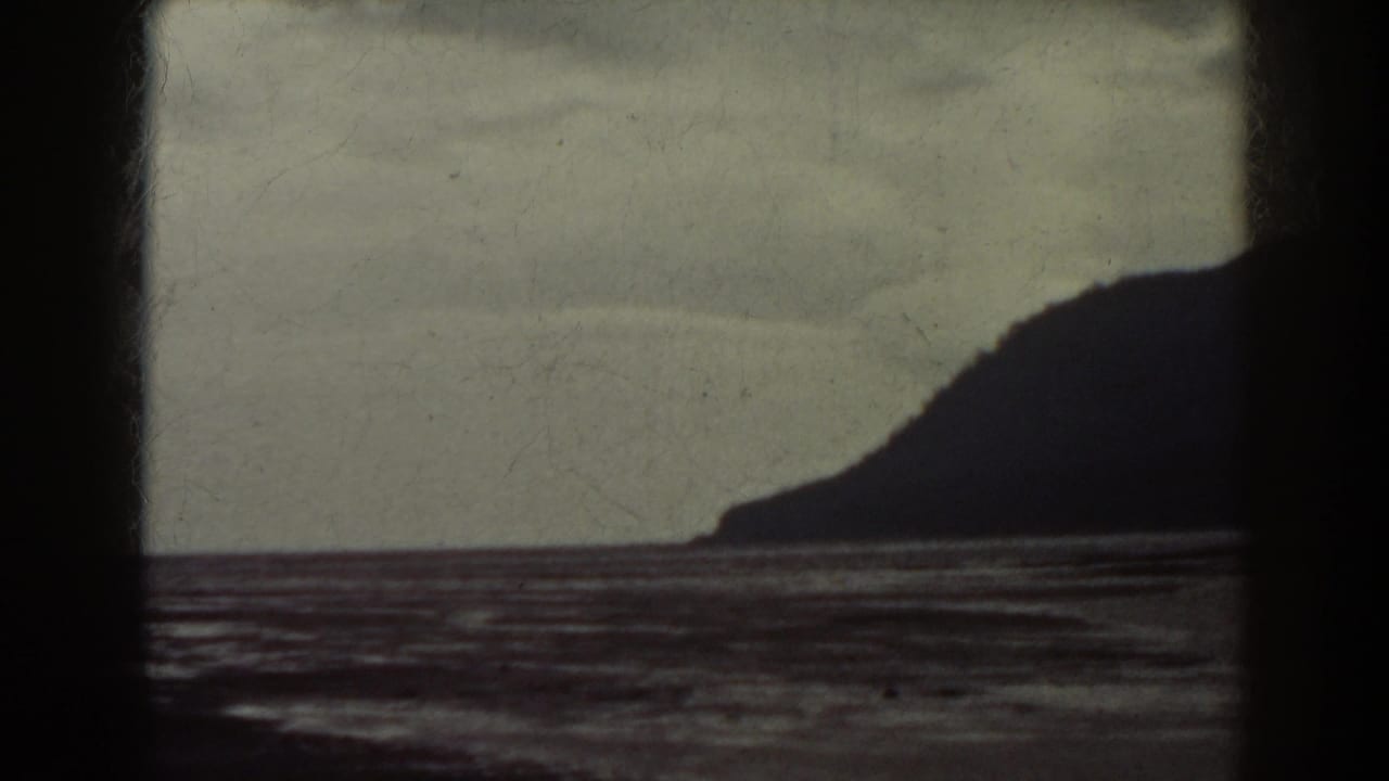1938 8mm frame with a dark landform silhouette on the right, rippled water below, and cloudy sky above.