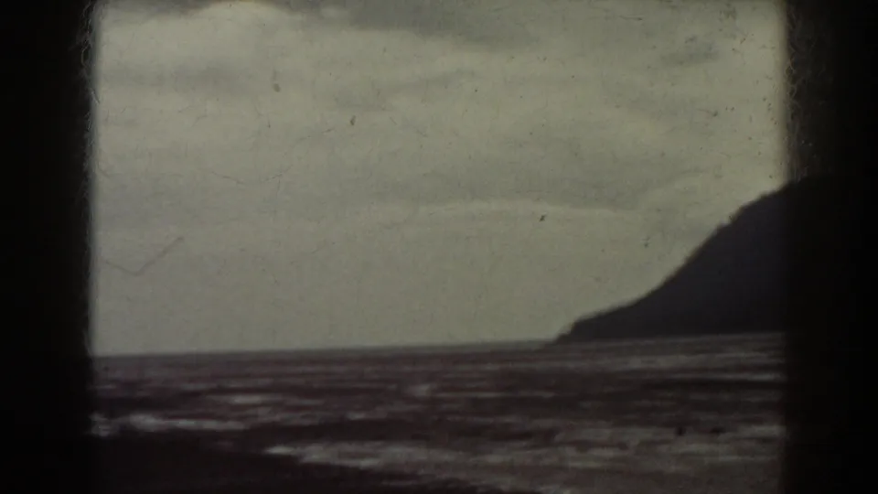 1938 8mm frame showing a dark slope beyond water under cloudy light, with contact-sheet and gate edges visible.