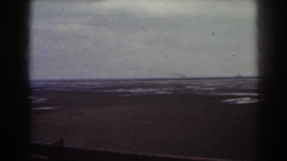 1938 8mm frame associated with Mount Washington, New Hampshire, showing a dark beach foreground and distant water beneath a muted sky.