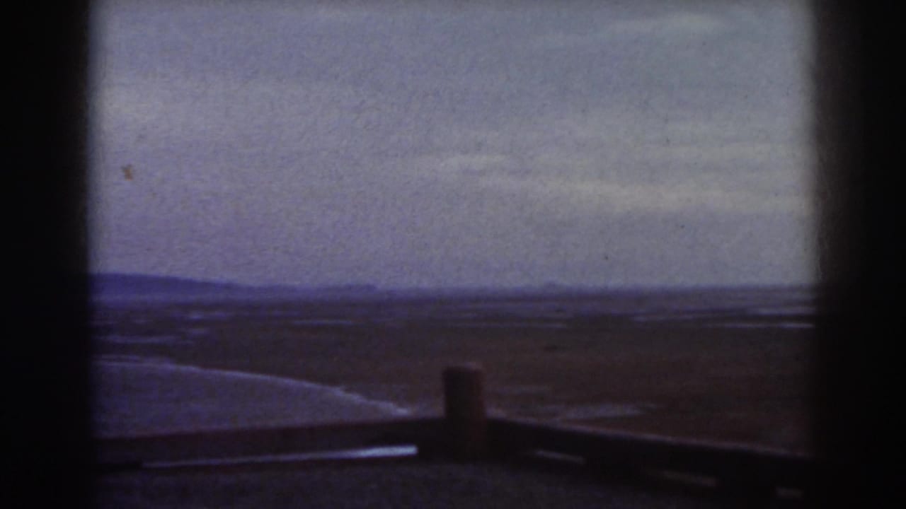 Dark-framed view across open terrain with a low barrier and distant hills under a pale sky.