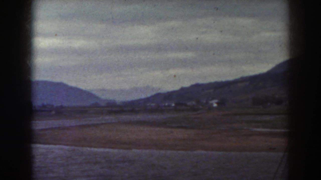 Brown field, water, hills, and cloudy sky framed by dark vertical edges.