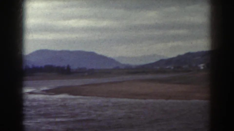 1938 home-movie frame of water and muddy shoreline with brown fields and hills in the distance near Mount Washington.