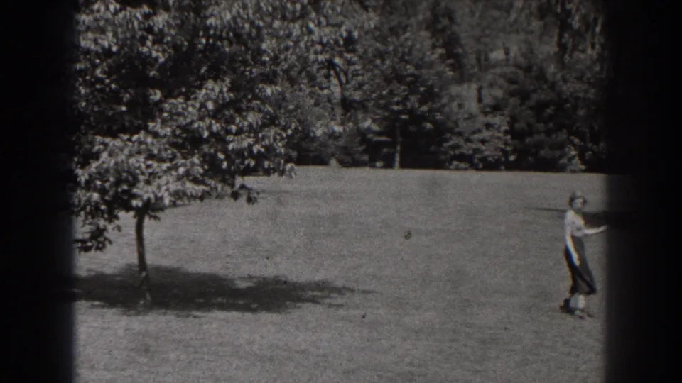 In 1938 New Jersey home-movie footage, a woman walks across a grassy park field near leafy trees.