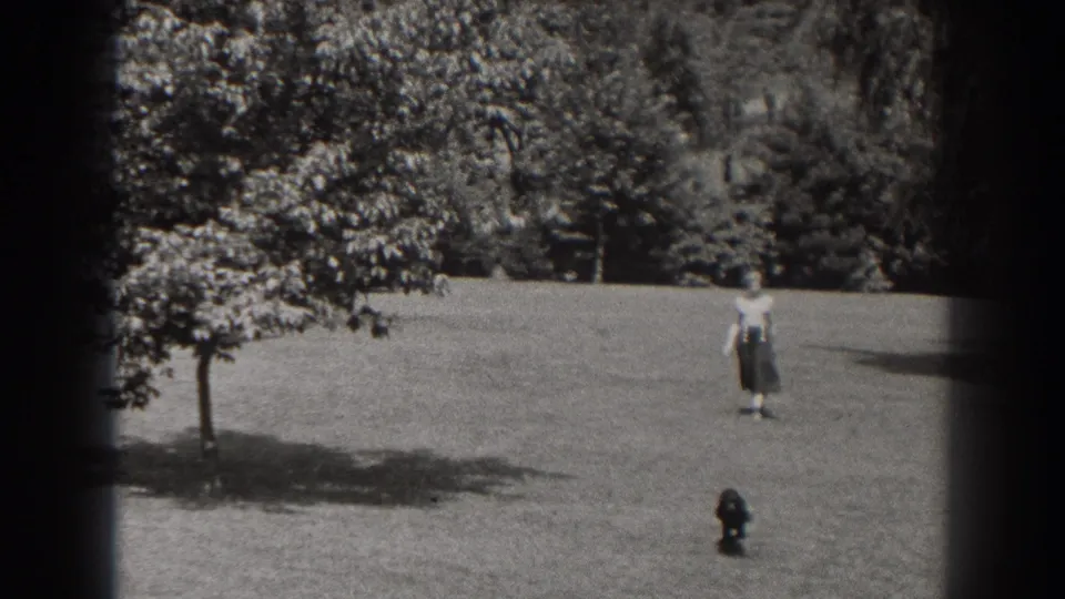 The small dark dog is closer to the camera while the woman remains farther back across the grassy field in black-and-white home-movie footage.