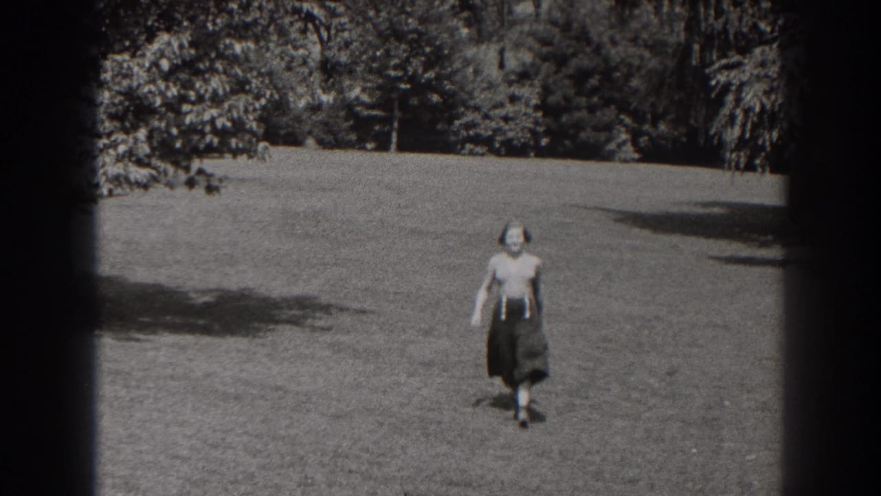Woman walking across a grassy lawn toward the camera with trees in the background.