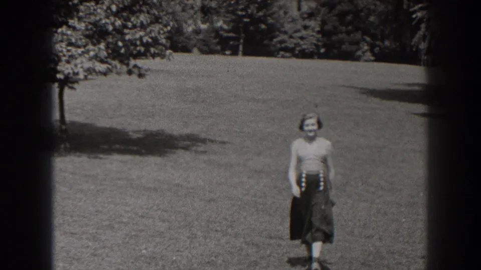 The woman smiles as she comes closer to the camera, framed by open grass and trees in the New Jersey home-movie shot.