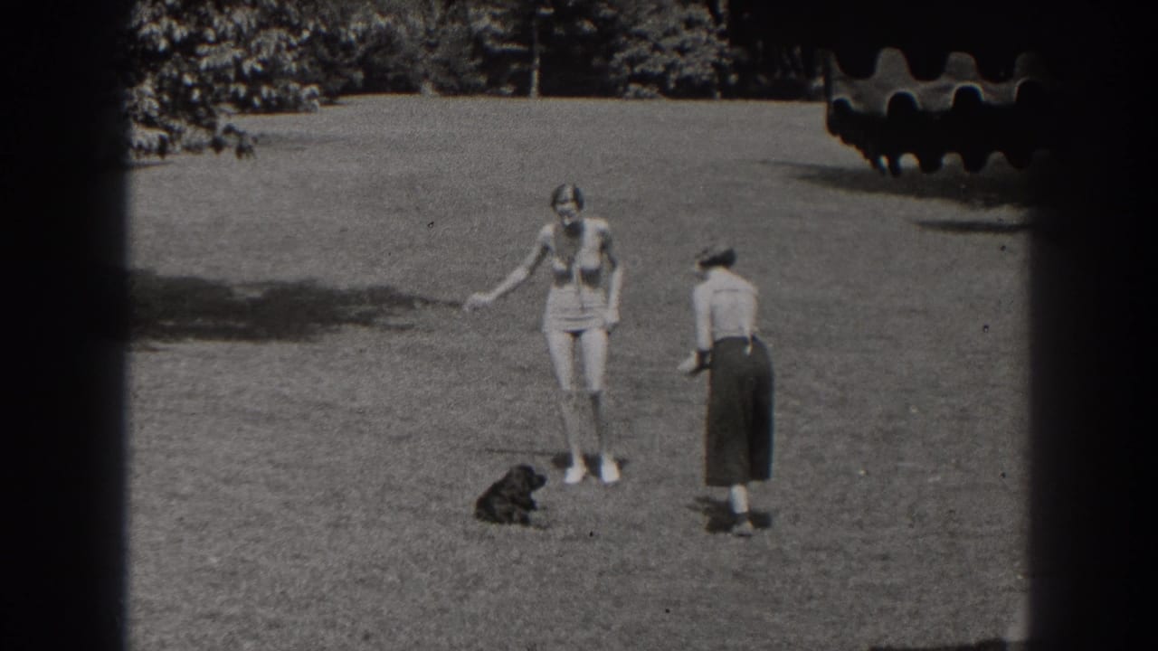 Two young women stand on a sunny lawn near a small black dog.