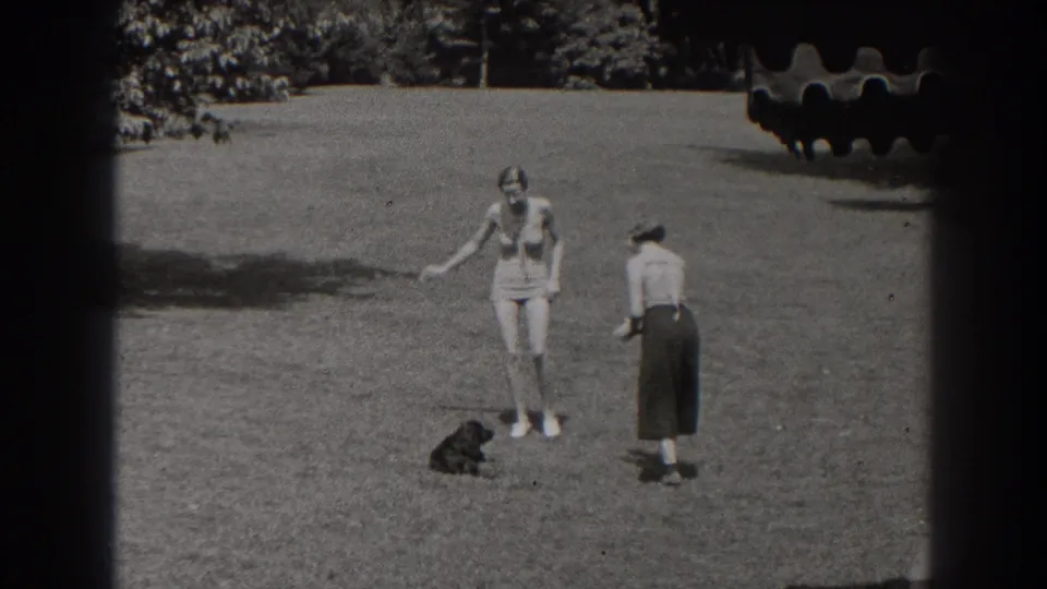 In 1938 New Jersey home-movie footage, a woman in a swimsuit gestures near a small black dog while another woman stands nearby on the lawn.