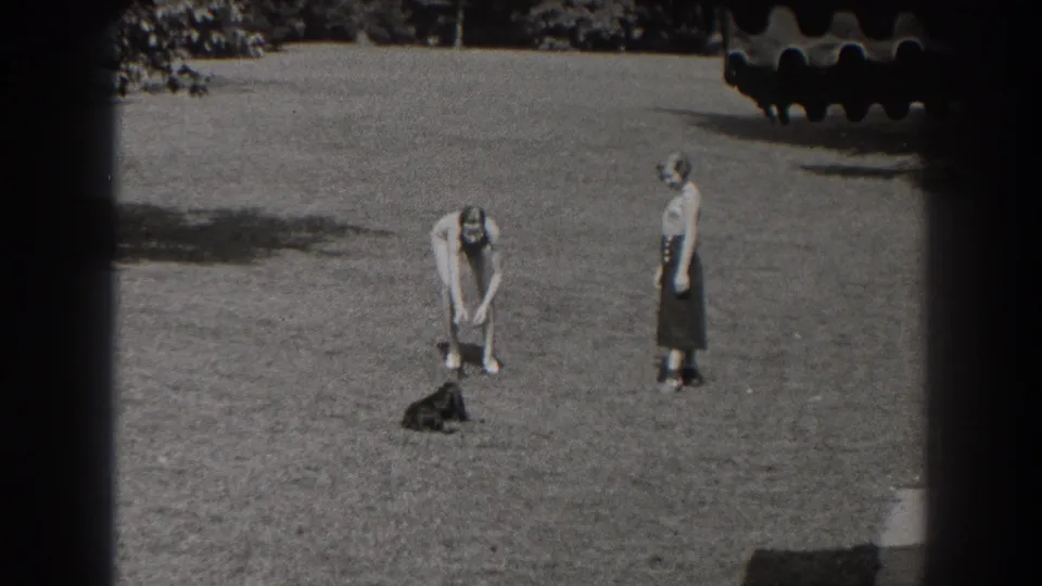 In the 1938 New Jersey yard scene, one young woman bends toward the small black dog as another woman in a dark skirt stands watching.