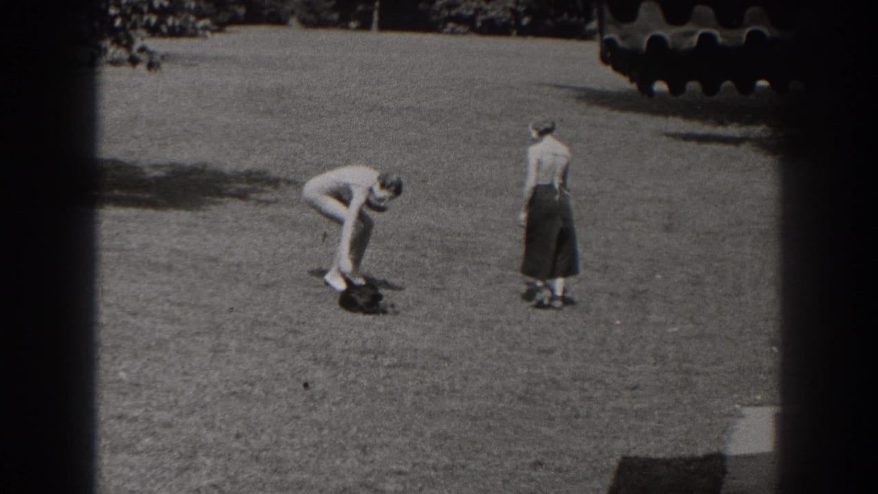 Man bends toward a small dark dog while a woman stands on a grassy lawn.