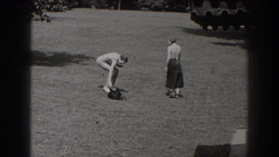 In 1938 New Jersey home-movie footage, a man in light clothing bends toward a small dark dog as a woman stands nearby on grass.