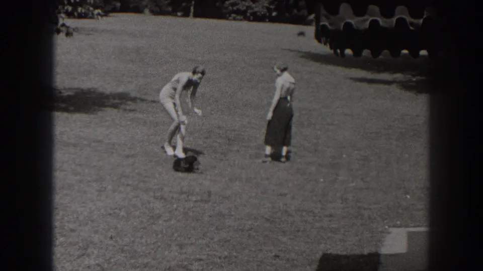 In the brief park sequence, the man rises near the small dark dog while the woman stands facing him across the grass.