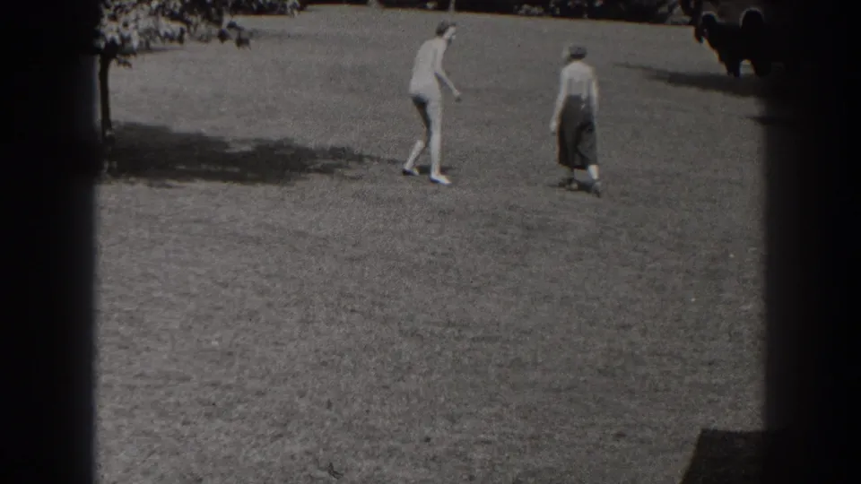 Two adults in 1930s attire stand or walk across a New Jersey lawn as a dog is visible near the frame edge.
