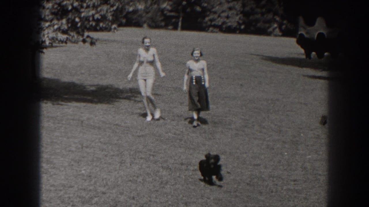 Two women walk across a grassy park as a small dark dog trots ahead.