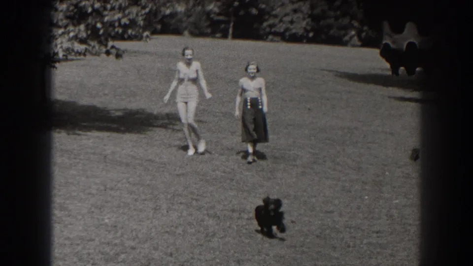 In 1938 New Jersey home-movie footage, two women cross a sunlit park lawn while a small dark dog moves in front of them.
