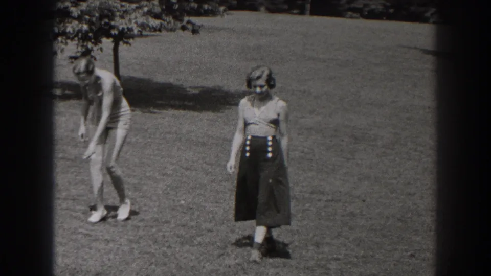 1938 New Jersey home-movie frame showing two women on a park lawn, one in a swimsuit bending down and one standing in a sleeveless top and dark skirt.