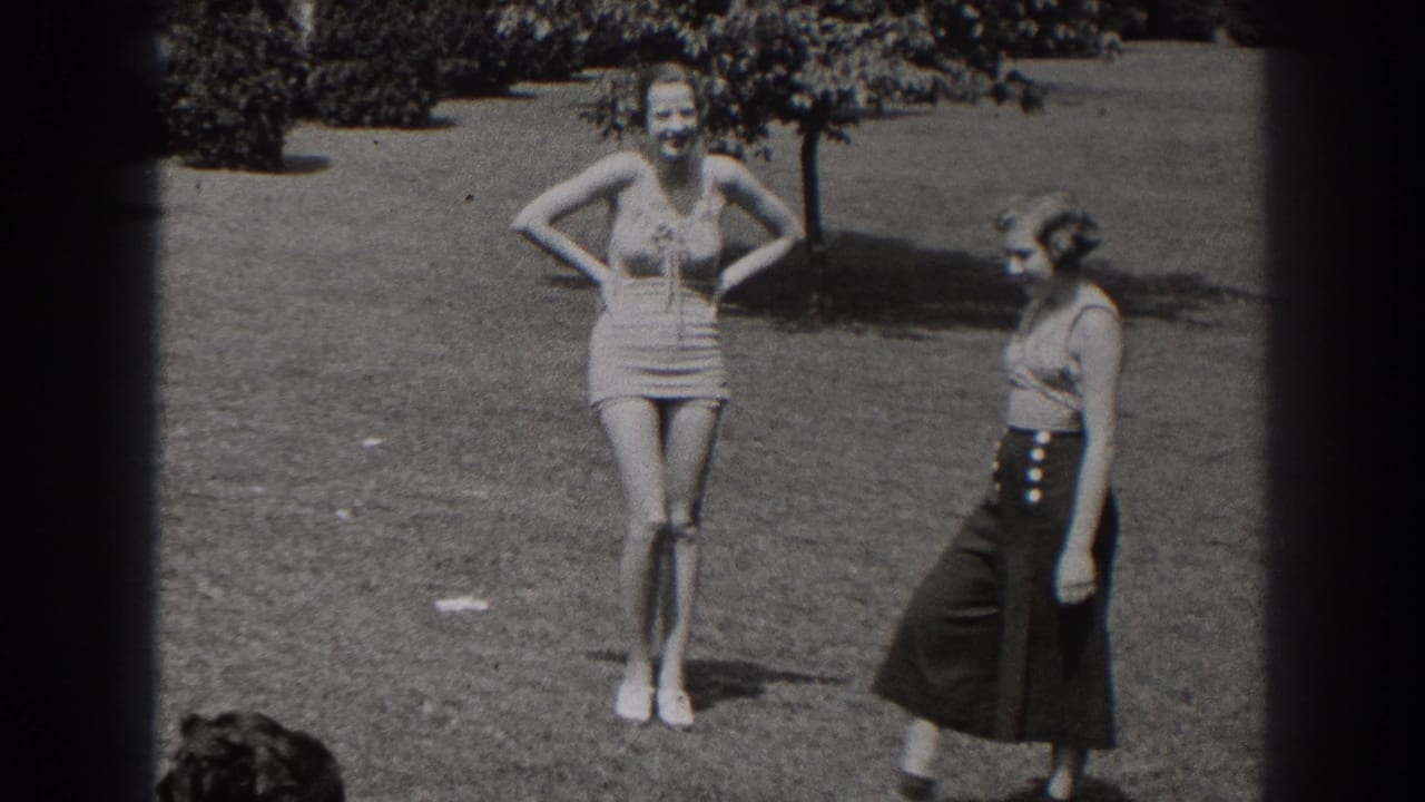 1938 New Jersey frame of two women on grass, with one smiling in a one-piece swimsuit and the other moving past in a sleeveless top and dark skirt.