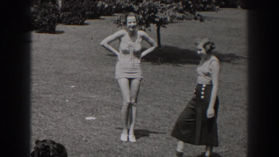1938 New Jersey frame of two women on grass, with one smiling in a one-piece swimsuit and the other moving past in a sleeveless top and dark skirt.