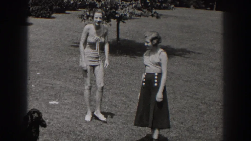 1938 New Jersey home-movie frame showing two women in summer clothing on a park lawn, with a small dark dog visible at the lower left.