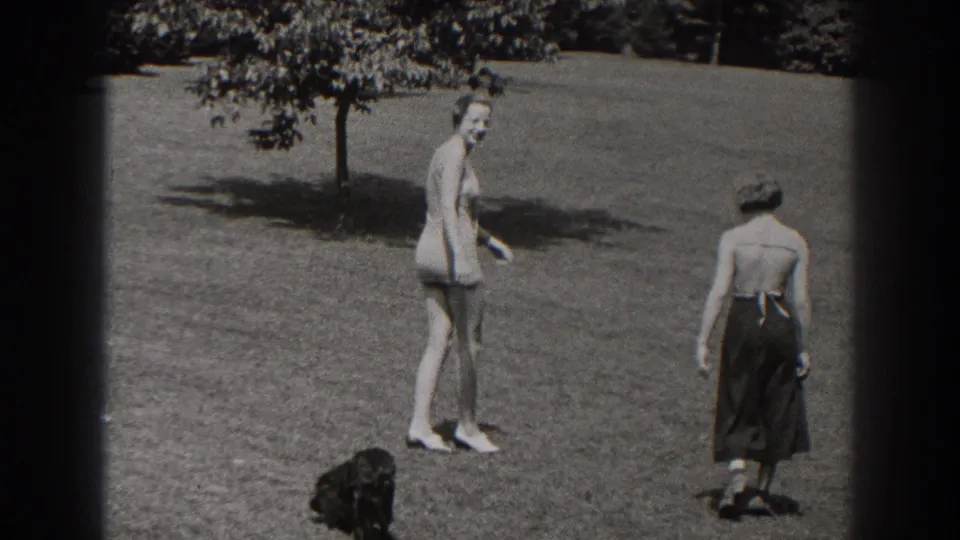 1938 New Jersey frame of two women moving across a grassy park area, one turning sideways in a swimsuit and one seen from behind in a dark skirt.