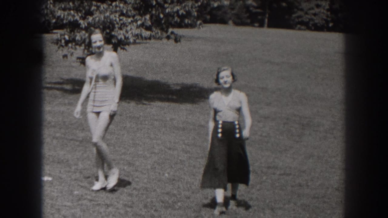 Two women stand and walk in a grassy field with trees behind them.