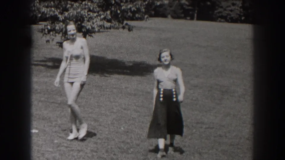 Two women in 1938 New Jersey home-movie footage walk across a sunny grassy field, one in a light fitted outfit and one in dark high-waisted pants.