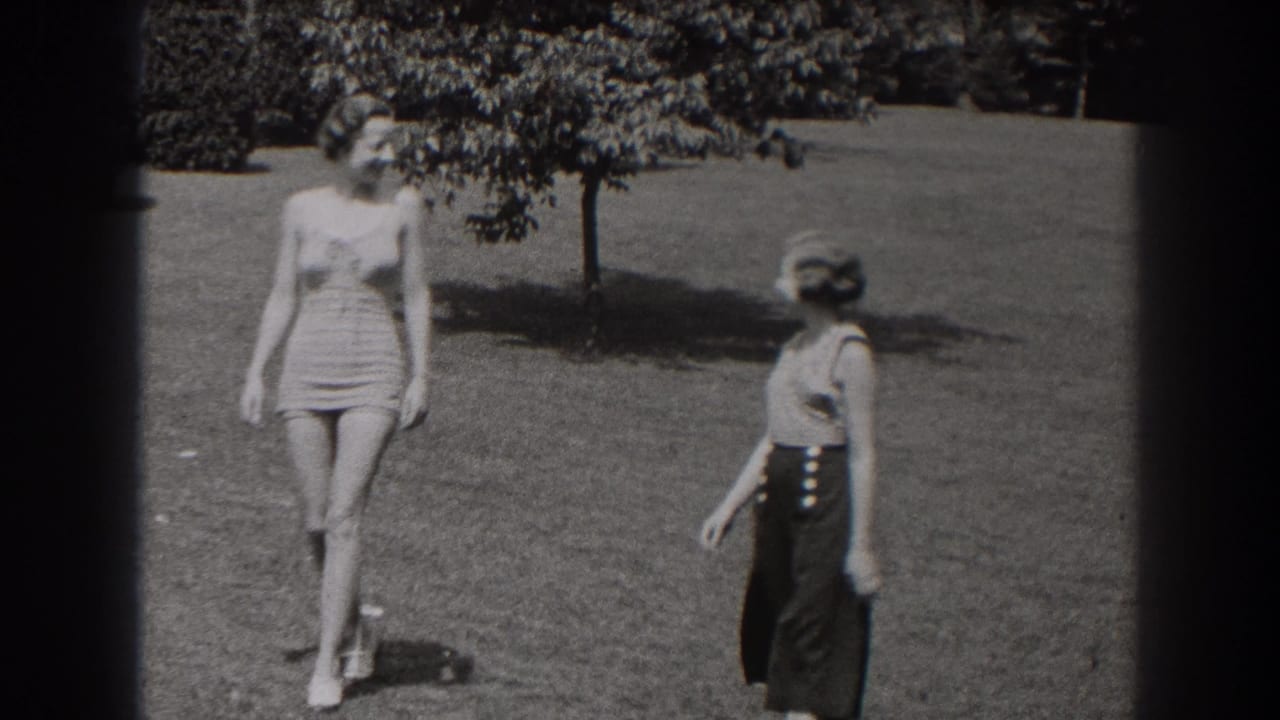 Two women cross a lawn, with one turning sideways near a tree-lined background.