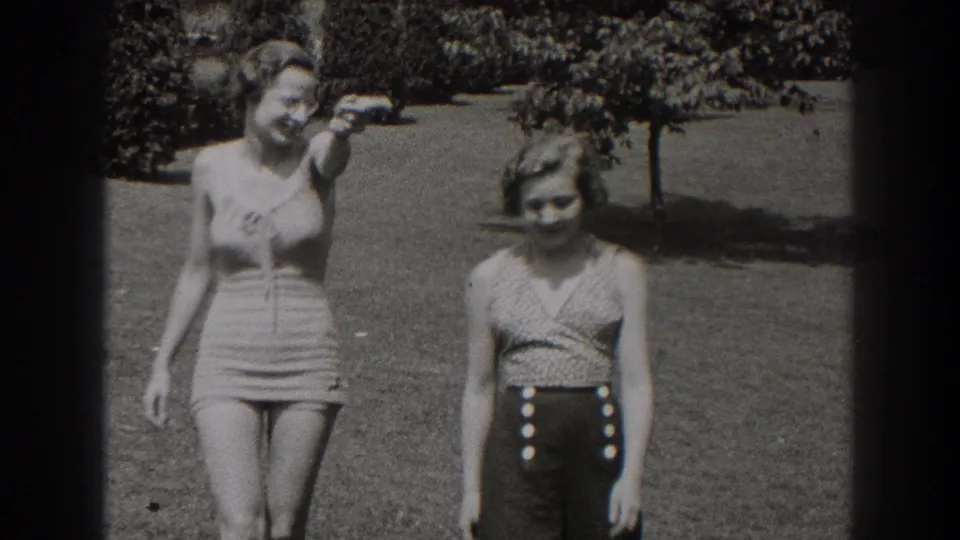 Two women in 1930s-style swimwear walk side by side across a sunny New Jersey lawn, with shrubs and trees behind them.