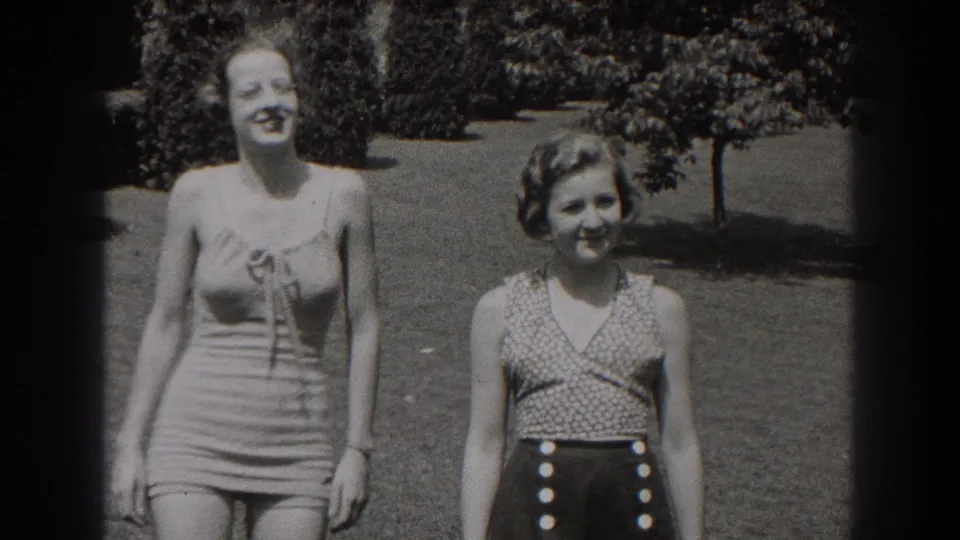 The two women face the camera in a sunny garden, one in a striped one-piece swimsuit and the other in a patterned top with high-waisted buttoned shorts.
