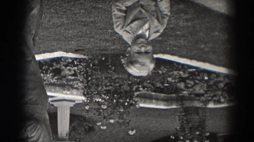 Late-1930s New Jersey 8mm still showing a reflected light-suited figure on a wet surface, framed by grass, walkway edges, and dark film borders.