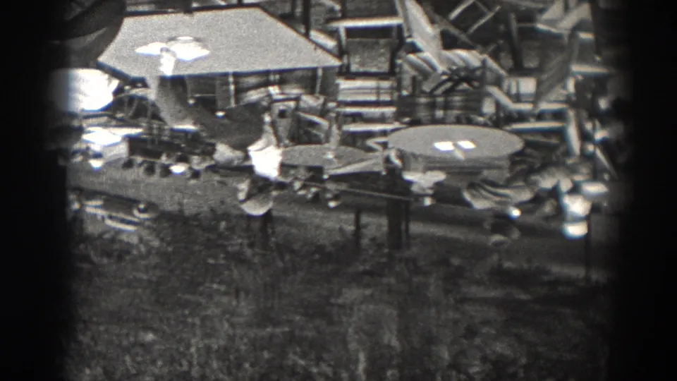 Black-and-white 8mm view of a restaurant or patio area with round tables, folding chairs, tableware, and adults partly visible at the edges.