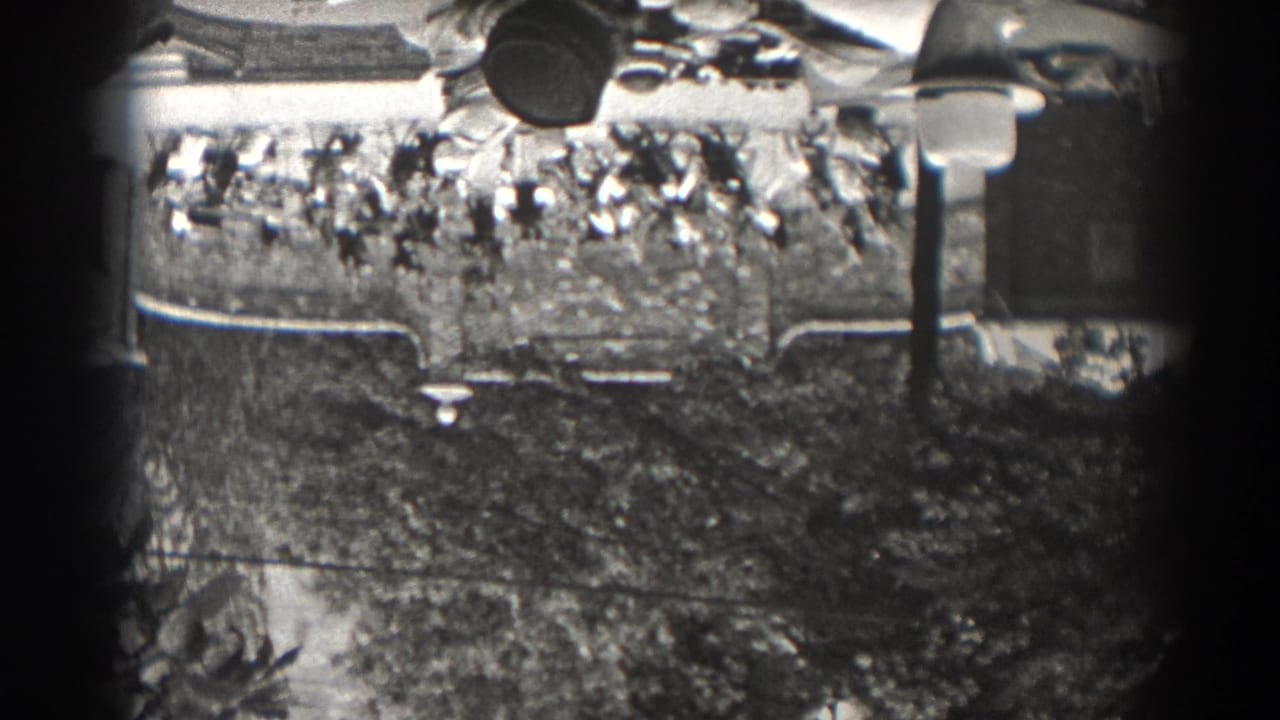 1938 New Jersey home-movie frame showing a distant seated crowd at an outdoor venue, partly obscured by trees and film-frame edges.