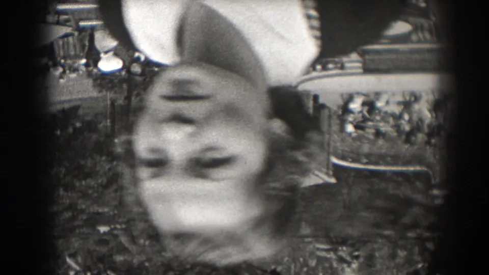 A blurred upside-down view shows a woman near a garden path in 1938 New Jersey, with foliage and a large building in the background.