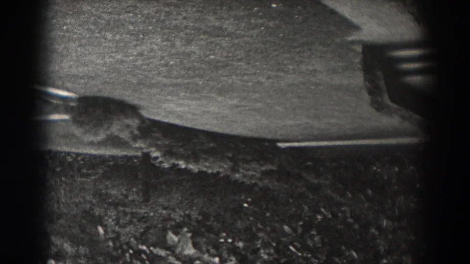 Upside-down black-and-white 8mm frame from New Jersey showing pavement, grass, and low shrubs under heavy vignette.