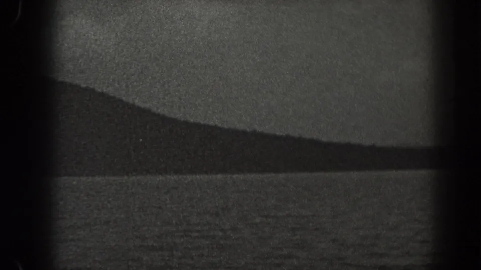 1938 New Jersey home-movie frame with calm water, a long dark ridge, and visible 8mm grain and gate shadowing.
