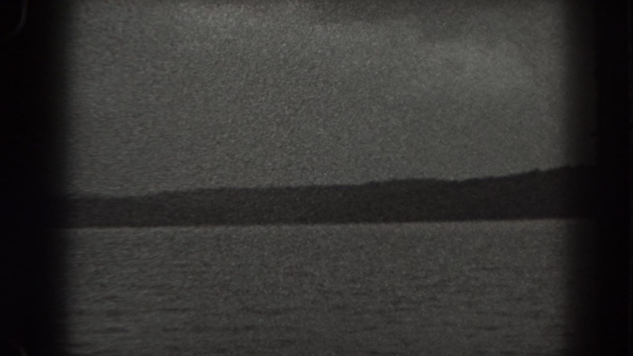 Rippling water below a distant dark wooded hill and muted sky.