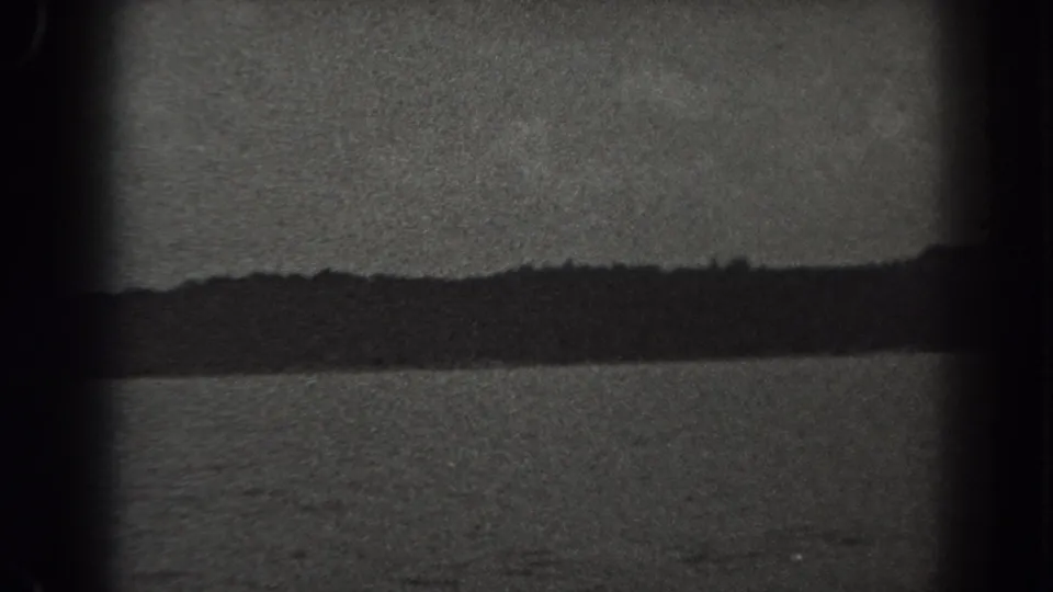 1938 New Jersey 8mm frame showing still lake water, a low dark shoreline, and overcast sky with strong film grain and edge shadowing.