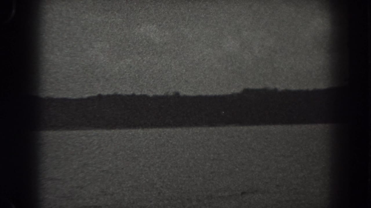 1938 New Jersey home-movie frame with a distant dark shoreline, gray sky, soft focus, grain, and visible gate shadows around the edges.