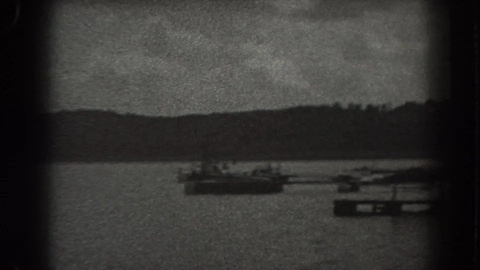 Grainy 1938 New Jersey 8mm frame with a boat, dock structures, open water, distant shoreline, and tiny indistinct figures.