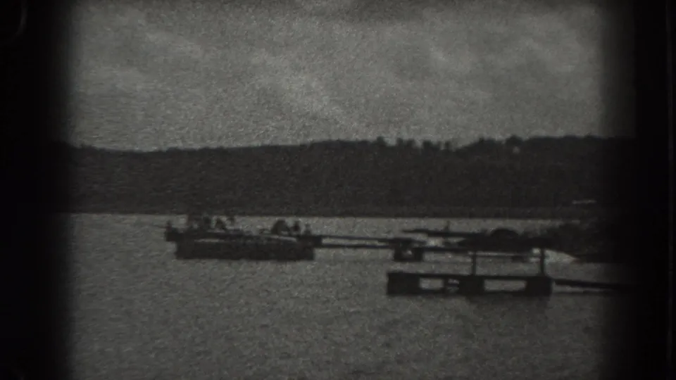 1938 New Jersey 8mm frame showing small boats, a dock or floating platform, lake water, and a distant tree-lined shore.