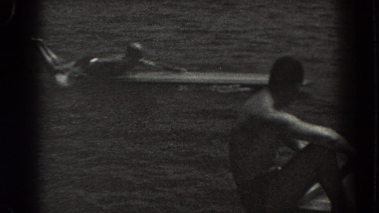 A man in dark shorts sits near the lake while another figure lies on a long board in the water.