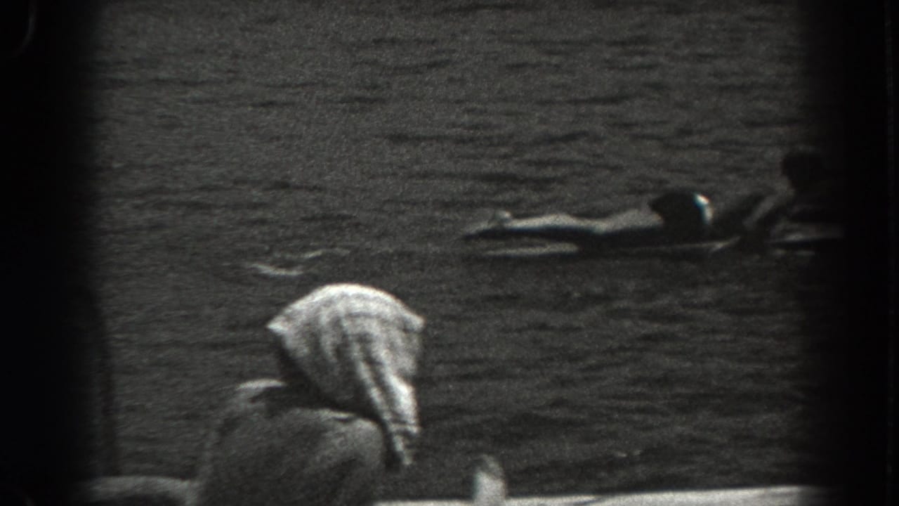 1938 New Jersey home-movie frame showing a dock edge, rippling water, and a person floating on a board beyond a foreground figure.