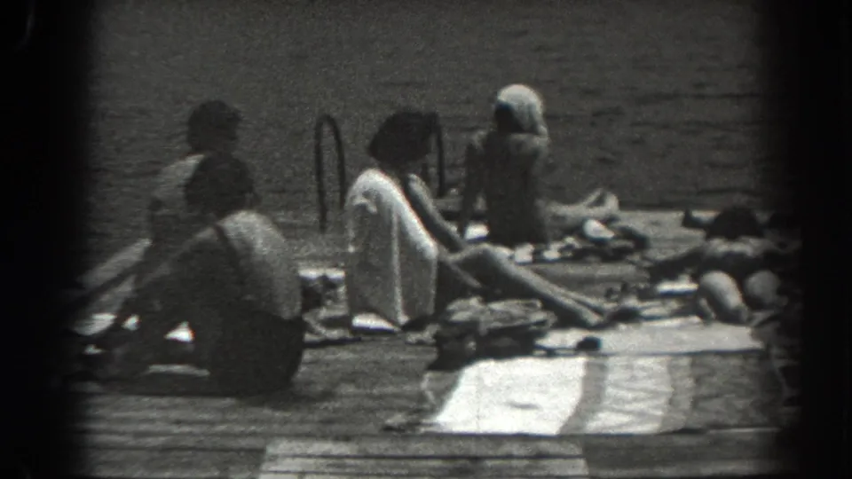 1938 New Jersey home-movie frame showing several people in swimwear resting on dock planks near water, towels, and a ladder.
