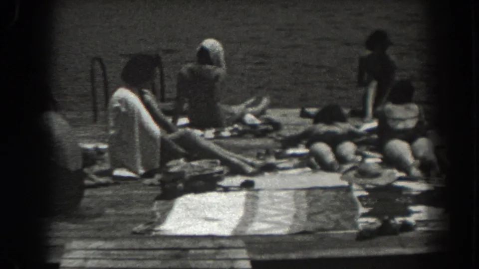 1938 New Jersey waterfront frame showing people on a wooden dock, some seated or lying down, with towels, belongings, and water behind them.